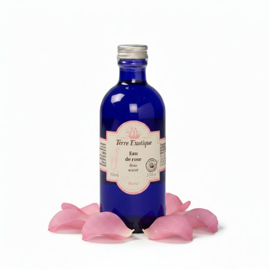A cobalt blue glass bottle of Terre Exotique Eau de rose base water with a silver cap and a pink and white label, set against a solid teal background.