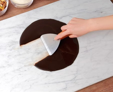 A marble stone working top being used for tempering chocolate. A person's hand is seen placing a piece of chocolate onto the marble surface.